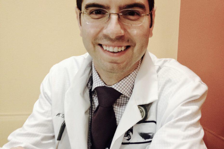 A man in a white doctor coat and stethoscope smiling.